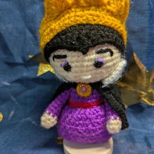 May include: Crocheted doll of a character wearing a yellow crown, black hair, and a purple dress with a red belt. The doll has a black cape and is standing on a wooden base. The doll's face has purple eye shadow and a stern expression.