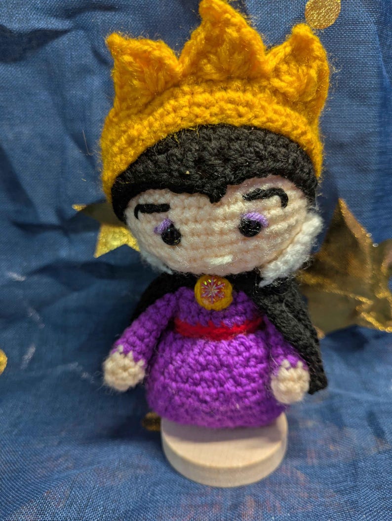 May include: Crocheted doll of a character wearing a yellow crown, black hair, and a purple dress with a red belt. The doll has a black cape and is standing on a wooden base. The doll's face has purple eye shadow and a stern expression.