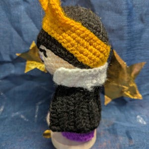 May include: Crocheted doll with a black head and body, a white collar, and a yellow crown. The doll is set against a blue background with gold star and circle accents. The doll's base is a light wood color.