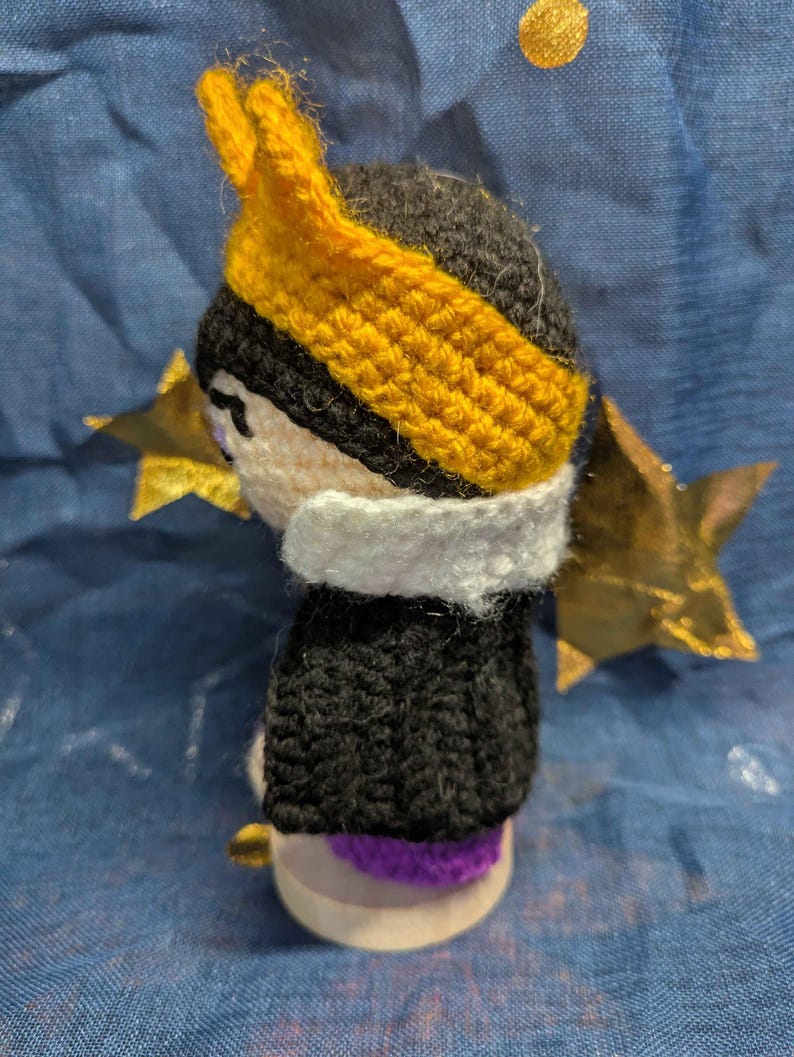 May include: Crocheted doll with a black head and body, a white collar, and a yellow crown. The doll is set against a blue background with gold star and circle accents. The doll's base is a light wood color.