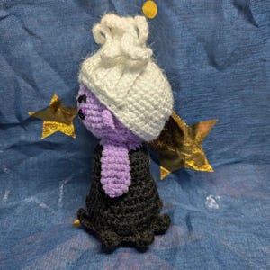 May include: A handmade crochet doll in the shape of a character with a purple face, a white hat, and a black dress. The doll has a purple neck piece and is set against a blue background with gold stars.