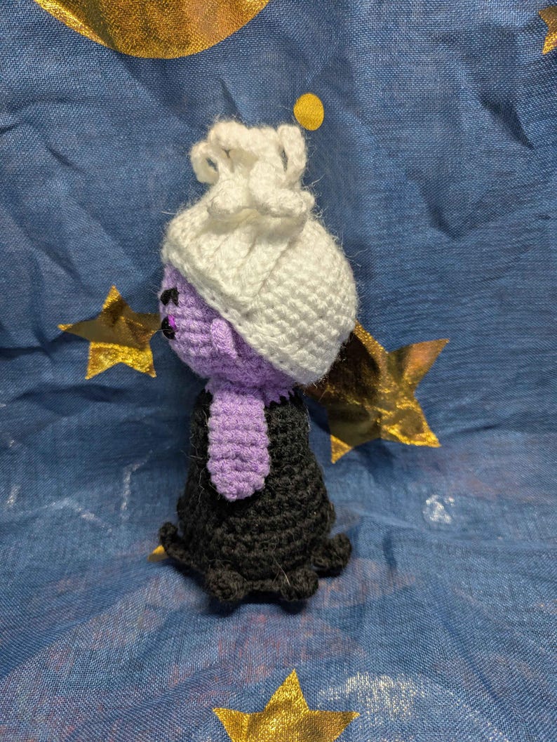 May include: A handmade crochet doll in the shape of a character with a purple face, a white hat, and a black dress. The doll has a purple neck piece and is set against a blue background with gold stars.
