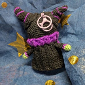 May include: A handmade black crochet doll with purple accents, including a collar, stripes on the horns, and a charm. The doll has green accents on the hands and is set against a blue fabric with gold star and dot embellishments.