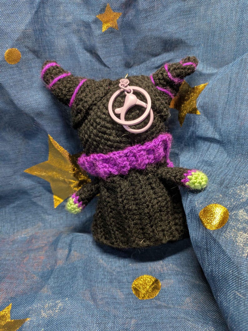 May include: A handmade black crochet doll with purple accents, including a collar, stripes on the horns, and a charm. The doll has green accents on the hands and is set against a blue fabric with gold star and dot embellishments.