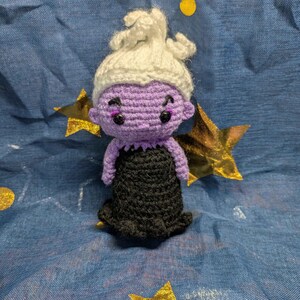 May include: A handmade crochet doll of a character with purple skin, a white updo, and a black dress. The doll has black eyes and is set against a blue background with gold stars.