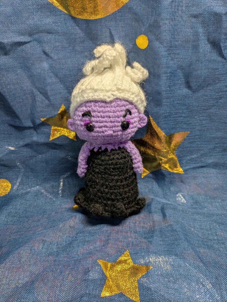 May include: A handmade crochet doll of a character with purple skin, a white updo, and a black dress. The doll has black eyes and is set against a blue background with gold stars.