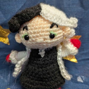 May include: Crocheted doll of a character with a black and white hairstyle, wearing a black dress, white coat, and red shoes. The doll has green eyes and a black beret. The doll is handmade and has a whimsical appearance.
