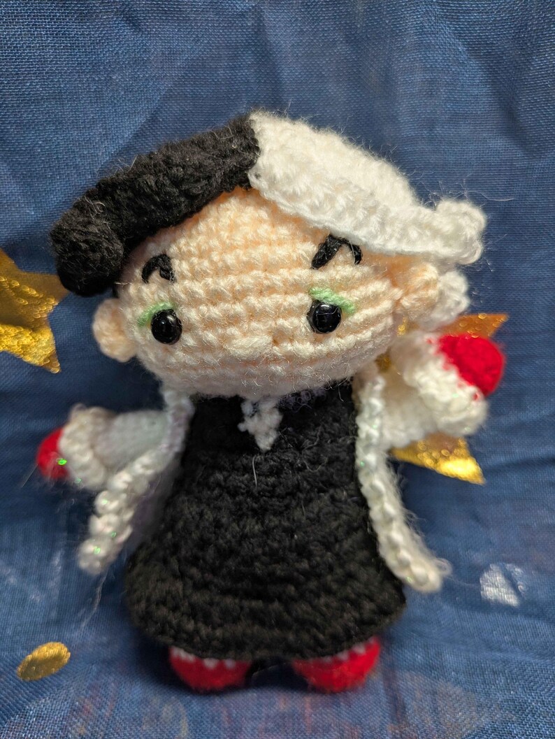 May include: Crocheted doll of a character with a black and white hairstyle, wearing a black dress, white coat, and red shoes. The doll has green eyes and a black beret. The doll is handmade and has a whimsical appearance.