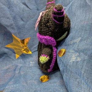 May include: A handmade black crochet cat with purple accents, including a scarf and stripes on the ears. The cat has green details on its paws. A small purple charm is attached. The background is blue with gold star and dot accents.