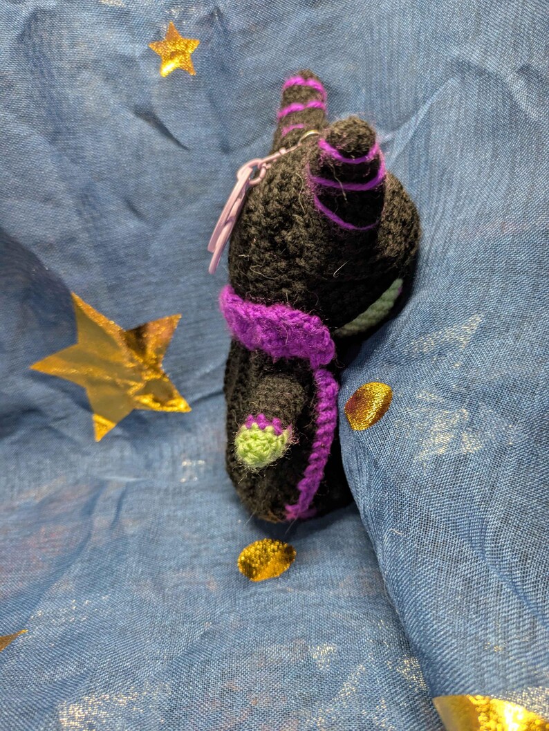 May include: A handmade black crochet cat with purple accents, including a scarf and stripes on the ears. The cat has green details on its paws. A small purple charm is attached. The background is blue with gold star and dot accents.