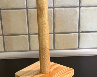 May include: A wooden paper towel holder. The base is a square of light-colored wood with visible grain. A vertical, cylindrical post rises from the center of the base. The holder is made of natural wood.