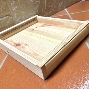 Puede incluir: Una bandeja cuadrada de madera sin terminar con una veta de madera clara y natural. La bandeja tiene un borde elevado y está construida con líneas simples y limpias. La madera parece ser pino o una madera blanda similar. La bandeja está sobre una superficie de baldosas de terracota.
