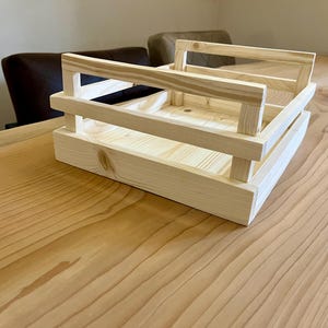 以下が含まれることがあります： 長方形のベースとハンドルが付いた、無垢材の木製クレート。未完成の木材で作られており、シンプルで素朴なデザインです。木製のテーブルの上に置かれています。