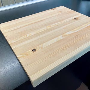 Può includere: Un tagliere rettangolare in legno chiaro con venature e nodi visibili. Il tagliere è composto da diverse assi di legno unite. Il tagliere è su una superficie scura.