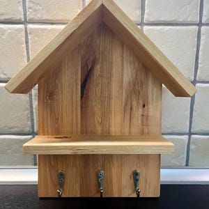 Può includere: Portachiavi in legno a forma di casa. Presenta un tetto a capanna, un piccolo ripiano e tre ganci in metallo per appendere le chiavi. Il legno ha una venatura naturale e un colore marrone chiaro.