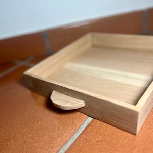 May include: A light-colored wooden tray with a rounded handle on one side. The tray is rectangular with a raised edge. The wood grain is visible. The tray is sitting on a brown tile surface.