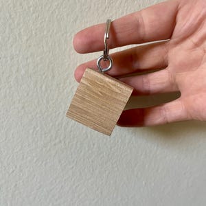 May include: A wooden cube keyring with a silver metal ring. The cube is made of light-coloured wood with visible grain. The keyring is held up by a hand against a neutral background.