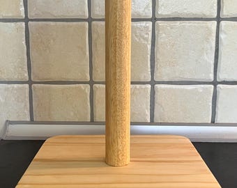 May include: A wooden paper towel holder. The base is a light-colored, rectangular block of wood with rounded corners. A vertical, cylindrical wooden dowel rises from the center of the base, designed to hold a roll of paper towels. The wood grain is visible.