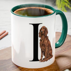 May include: White ceramic mug with a green handle and rim, featuring a black letter "I" and a brown dog illustration. The mug is filled with a dark liquid and sits on a wooden cutting board with cinnamon sticks and a pine cone.