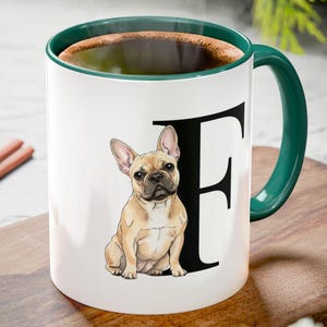 Può includere: Tazza in ceramica bianca con manico e bordo verdi, con un'illustrazione di un bulldog francese marrone e la lettera "F". La tazza è riempita con un liquido scuro, forse caffè o tè, e poggia su un tagliere di legno.