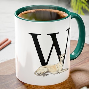 May include: White ceramic mug with a green handle and interior, featuring a large black letter "W" and a drawing of a greyhound dog. The mug is filled with a dark liquid, possibly coffee or tea.