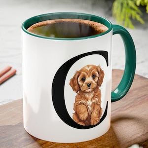 May include: White ceramic mug with a green handle and rim, featuring a brown puppy illustration inside a black letter "C". The mug is filled with a dark liquid and sits on a wooden cutting board with cinnamon sticks and pine cones.