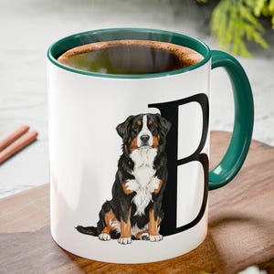 Könnte beinhalten: Weißer Keramikbecher mit grünem Henkel und Rand, mit einem schwarzen Buchstaben "B" und einer Illustration eines Berner Sennenhundes. Der Becher ist mit einer dunklen Flüssigkeit gefüllt und steht auf einem Holzschneidebrett.