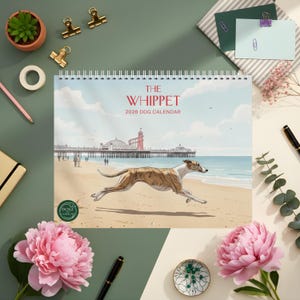 May include: A 2026 dog calendar featuring a running Whippet on a beach scene. The calendar has a spiral binding and the text "THE WHIPPET" in red. The image includes a pier, ocean, and a sandy beach. The calendar is surrounded by office supplies and flowers.