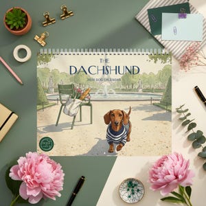 May include: A spiral-bound 2026 calendar titled "The Dachshund" featuring an illustration of a dachshund wearing a blue and white striped sweater. The calendar's background depicts a park scene with a fountain and a chair with a bag of groceries.