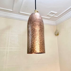May include: A copper-coloured pendant light fitting with a hammered texture. The light hangs from a black cord and has a brass fitting at the top. The fitting is cylindrical with a rounded top.