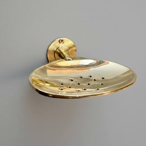Close-up of a handmade solid brass wall-mounted soap dish with drainage holes and polished living finish.