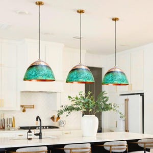 Op de afbeelding: Drie hanglampen met een koperkleurige rand en een groen en blauw getextureerde koepel. De lampen hangen aan een messingkleurige plafondbevestiging. De lampen bevinden zich in een keuken met witte kasten en een wit aanrecht.