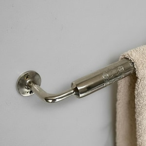 May include: A silver-toned towel bar mounted on a light gray wall. The bar has a decorative, patterned design on the end that holds the towel. A beige towel is draped over the bar.