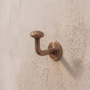 May include: A wall-mounted hook with a rounded knob end and a circular base plate. The hardware features a weathered, metallic copper finish with visible dark spots and a textured surface, secured to the wall with a single visible screw.