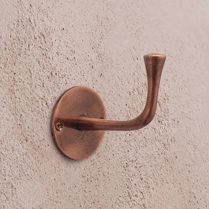 Könnte beinhalten: Ein Wandhaken mit metallischem Kupfer-Finish. Der Haken verfügt über eine kreisförmige Grundplatte, die mit einer sichtbaren Schraube befestigt ist, und einen geschwungenen Arm, der nach außen ragt und in einer leicht ausgestellten Spitze endet.