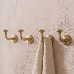 Può includere: Quattro ganci da parete in ottone con finitura lucida, ciascuno dotato di una base circolare e un braccio curvo con punta arrotondata. Un gancio sostiene un asciugamano in tessuto color bianco sporco con motivo a spina di pesce su una parete.