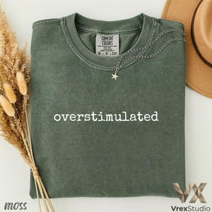 以下が含まれることがあります： 「overstimulated」の白い文字がプリントされたモスグリーンのTシャツ。Tシャツはクルーネックと半袖です。星のペンダントが付いたシルバーのネックレスがTシャツの上にかけられています。茶色の帽子が背景に見えます。