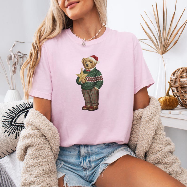 May include: A light pink t-shirt featuring a graphic of a teddy bear wearing a Santa hat, green sweater, and plaid pants, holding a gold star. The bear is a festive design for the holiday season.