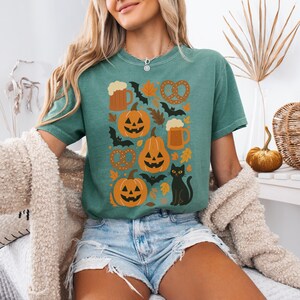 May include: Teal green t-shirt featuring a Halloween-themed design. The graphic includes orange jack-o'-lanterns, bats, pretzels, beer mugs, and autumn leaves. A black cat is also featured in the design. The shirt is a comfortable, casual style.