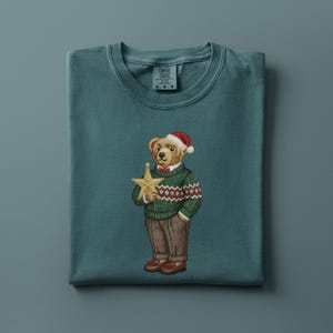 May include: Teal-colored t-shirt featuring an embroidered bear wearing a Santa hat, green sweater with a red and white pattern, and brown pants. The bear holds a gold star. The shirt has a crew neck and the brand name is visible on the upper left.
