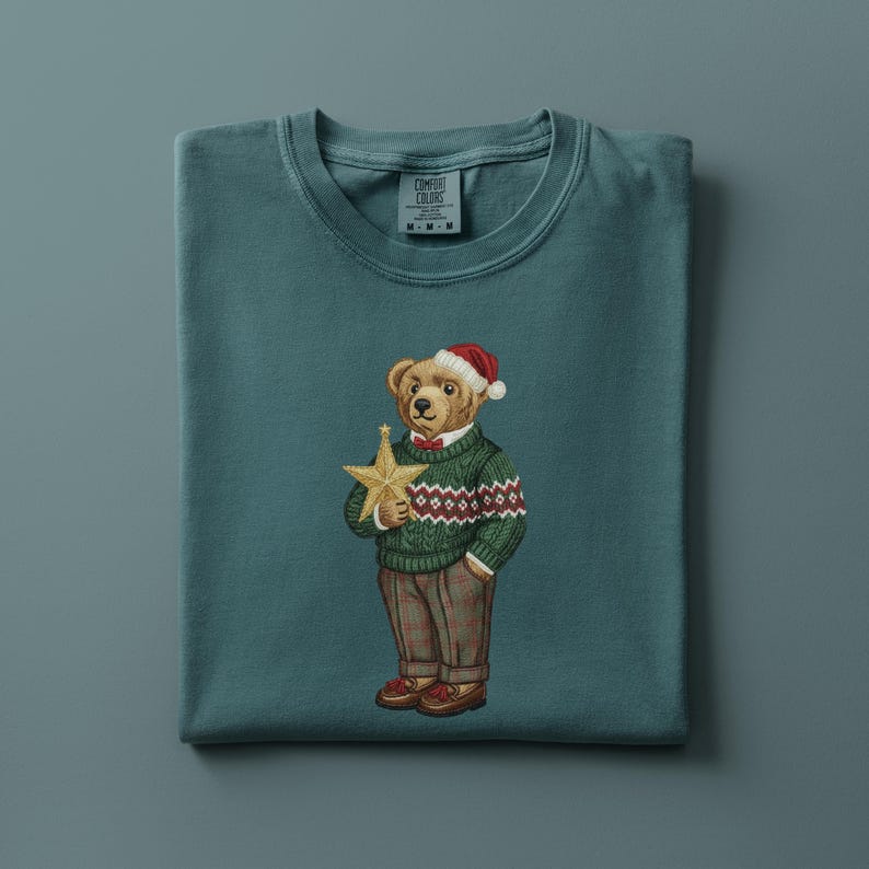May include: Teal-colored t-shirt featuring an embroidered bear wearing a Santa hat, green sweater with a red and white pattern, and brown pants. The bear holds a gold star. The shirt has a crew neck and the brand name is visible on the upper left.