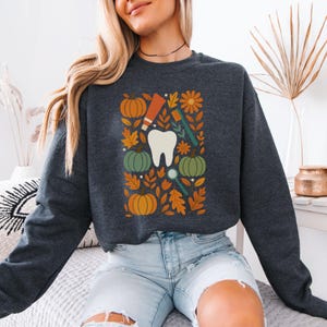 May include: Dark gray sweatshirt featuring a dental-themed design. The graphic includes a large white tooth, pumpkins, fall leaves, a toothbrush, toothpaste, and a dental mirror. The design is in shades of orange, green, and brown.
