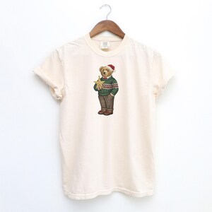 May include: Cream-colored t-shirt featuring a detailed illustration of a bear wearing a green sweater with a red and white pattern, brown pants, and a Santa hat. The bear is holding a gold star. The shirt is hanging on a wooden hanger.