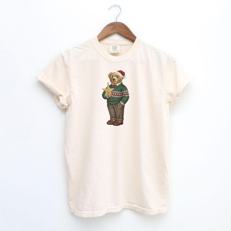 May include: Cream-colored t-shirt featuring a detailed illustration of a bear wearing a green sweater with a red and white pattern, brown pants, and a Santa hat. The bear is holding a gold star. The shirt is hanging on a wooden hanger.