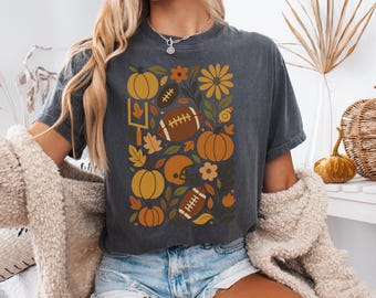 May include: Dark grey t-shirt featuring a fall-themed design. The graphic includes pumpkins, footballs, leaves, and flowers in shades of orange, yellow, and brown. A football helmet and goal post are also included in the design.