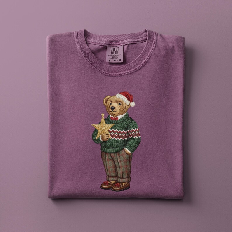 May include: A purple Comfort Colors t-shirt featuring an embroidered bear wearing a Santa hat and a green sweater with a red and white pattern. The bear holds a gold star. The shirt is folded neatly.