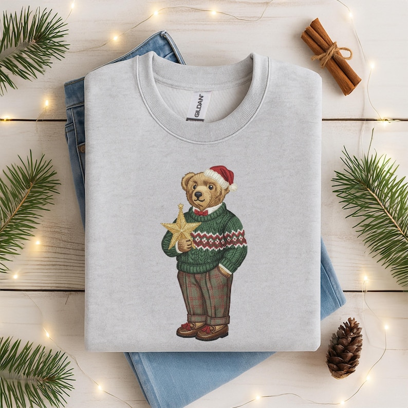 May include: A light gray sweatshirt featuring an embroidered bear wearing a Santa hat, green sweater with a red and white pattern, and plaid pants. The bear is holding a gold star. A festive holiday design.