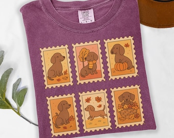 May include: A purple Comfort Colors t-shirt featuring six postage stamp designs with brown dachshund illustrations. The stamps have autumn-themed backgrounds with leaves and pumpkins. The shirt has a crew neck and short sleeves.