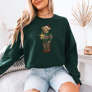 May include: Dark green sweatshirt featuring an embroidered bear wearing a Santa hat, sweater, and pants, holding a gold star. The bear design has a red bow tie and brown shoes. The sweatshirt is a solid color.
