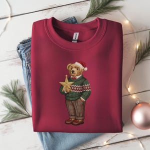 May include: Burgundy crewneck sweatshirt featuring an embroidered bear wearing a Santa hat, green sweater, and plaid pants. The bear holds a gold star. A festive holiday design.